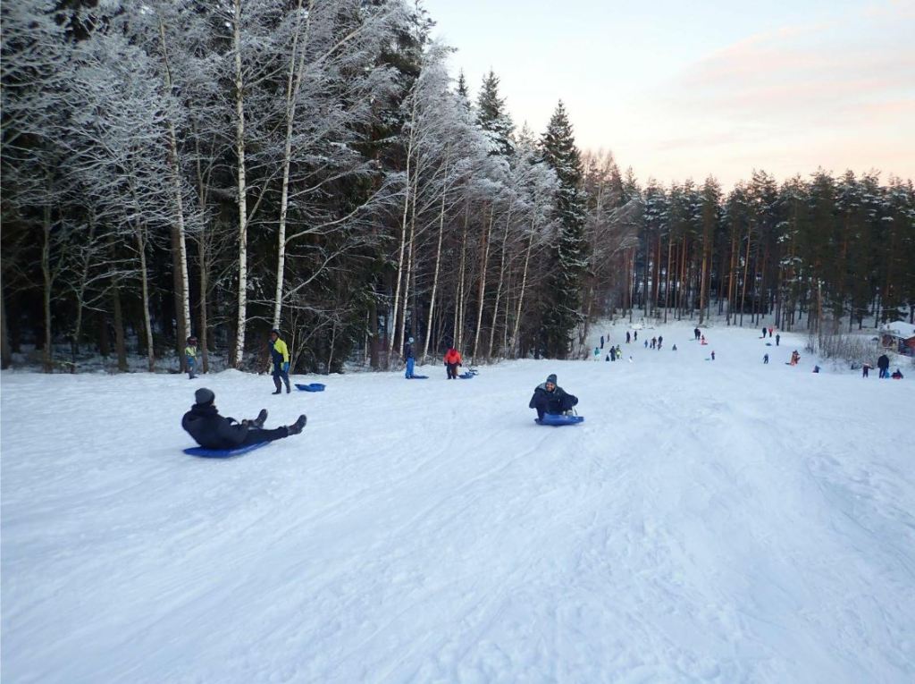 People sliding down a snow-covered hill. Trees on the side and the bottom of the hill. Paradisbacken in Borlänge.